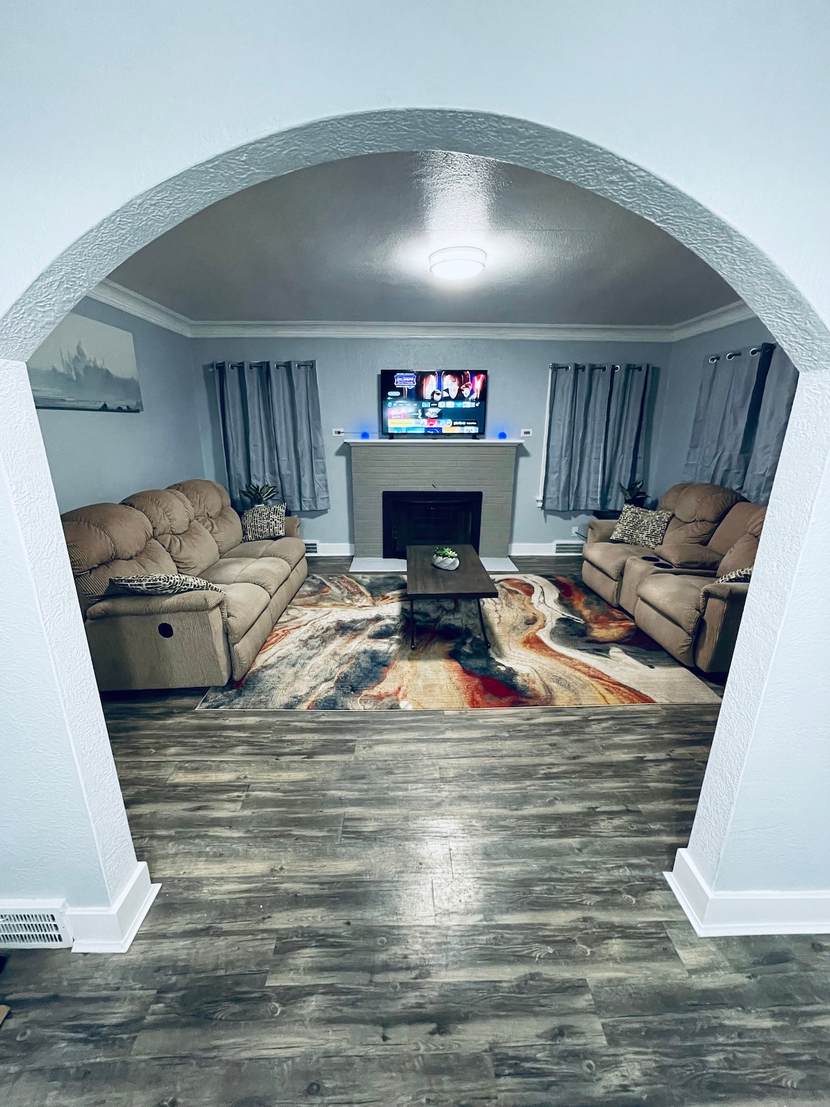 The living room is framed by an arched entryway, featuring two comfortable sofas arranged around a central coffee table. A large area rug adds warmth to the space, and a television is mounted above the fireplace, flanked by light-filtering curtains.