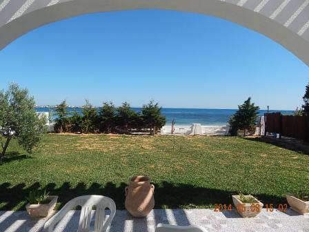 An expansive lawn is visible under a clear blue sky, bordered by lush greenery and leading to the ocean. Two potted plants sit on the ground near white chairs, while the shoreline can be seen in the background, providing a serene coastal view.