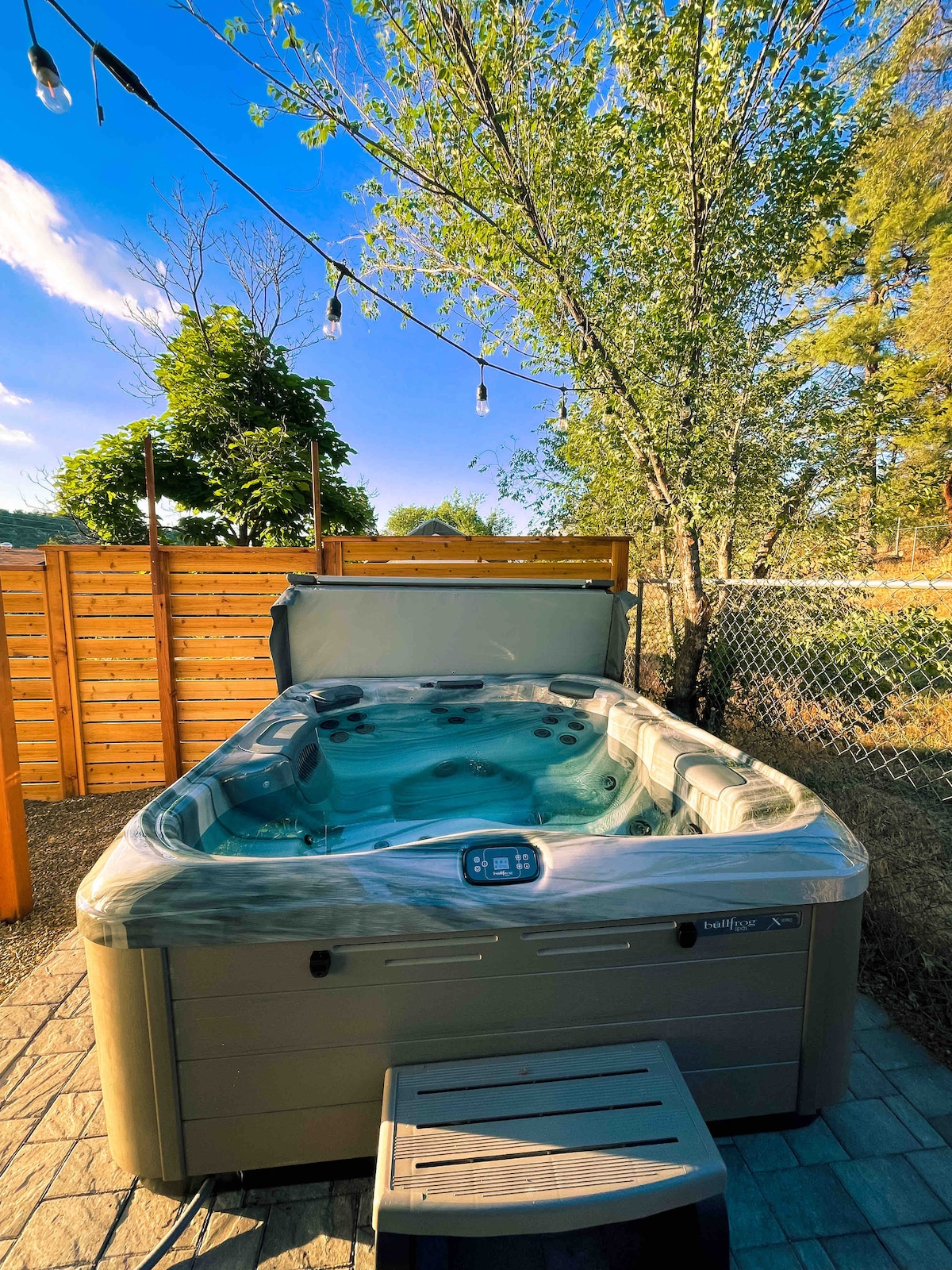 A private hot tub is positioned on a stone-paved area, surrounded by wooden fencing and greenery. The tub's cover is partially open, revealing the inviting water inside. Decorative string lights are draped above, contributing to the peaceful ambiance of the outdoor space.