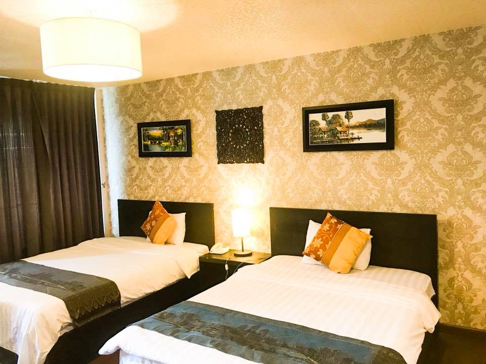 The room features two spacious beds with elegant linens and decorative pillows. A bedside lamp provides soft illumination. Artwork hangs on the patterned wallpaper, and a window is covered by dark curtains, allowing for privacy while providing a cozy ambiance.