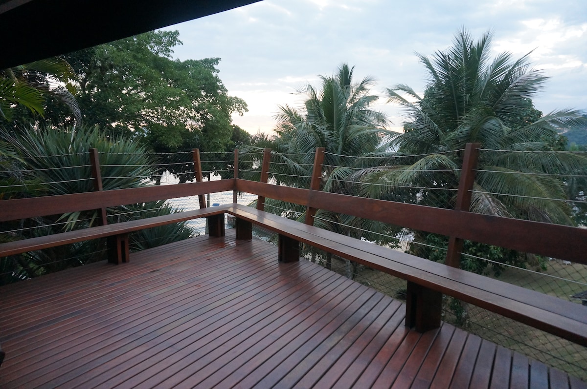 A wooden deck is seen, featuring long benches along the perimeter. Lush green trees and palm trees frame the view, while the ocean is visible beyond. The sky is lightly overcast, adding a serene ambiance.
