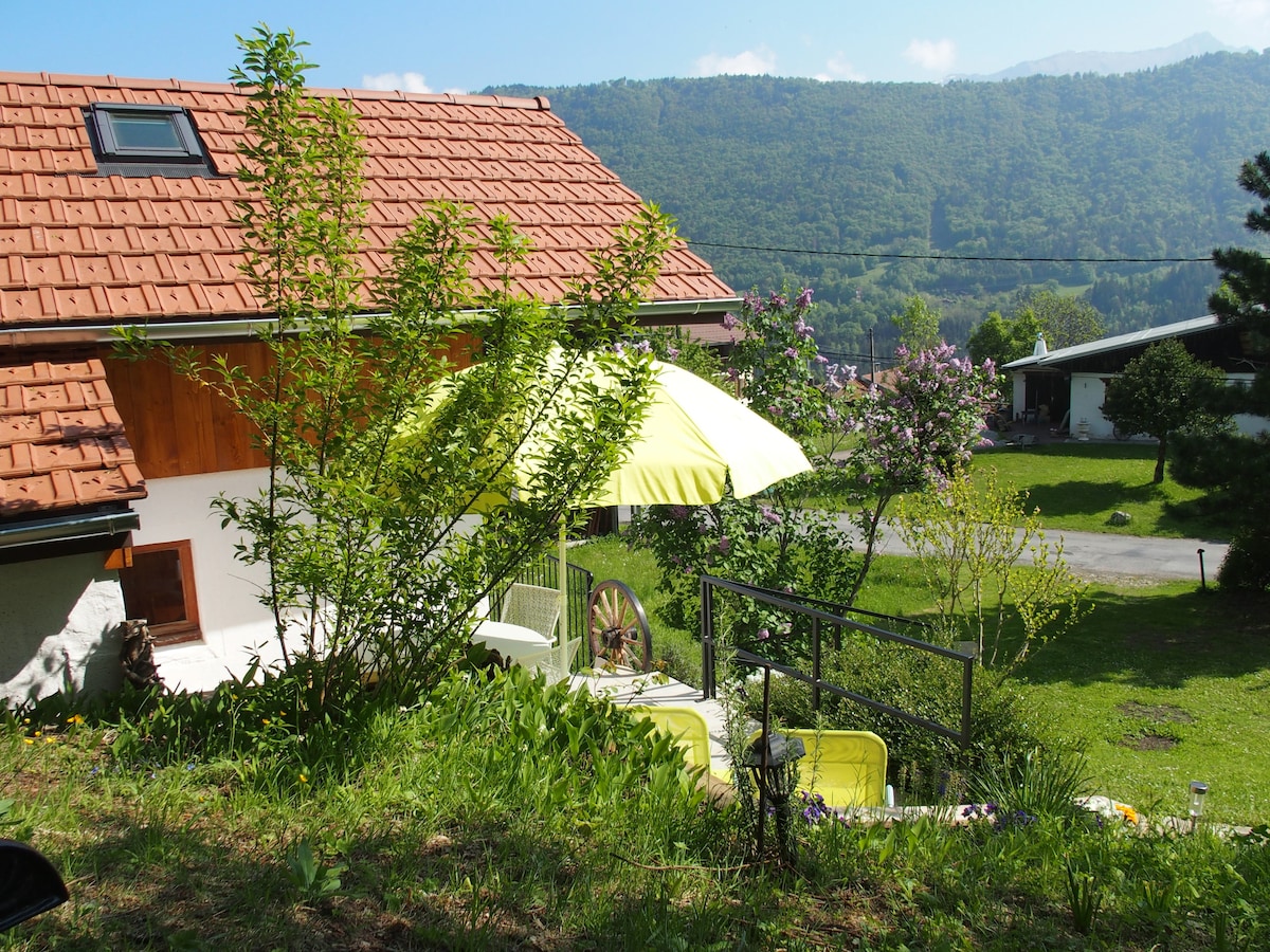 A quaint independent house is set against a backdrop of green hills. A yellow umbrella provides shade over outdoor seating, surrounded by lush grass and blooming flowers. The structure features a sloped roof with a skylight and is complemented by a pathway leading to the entrance.