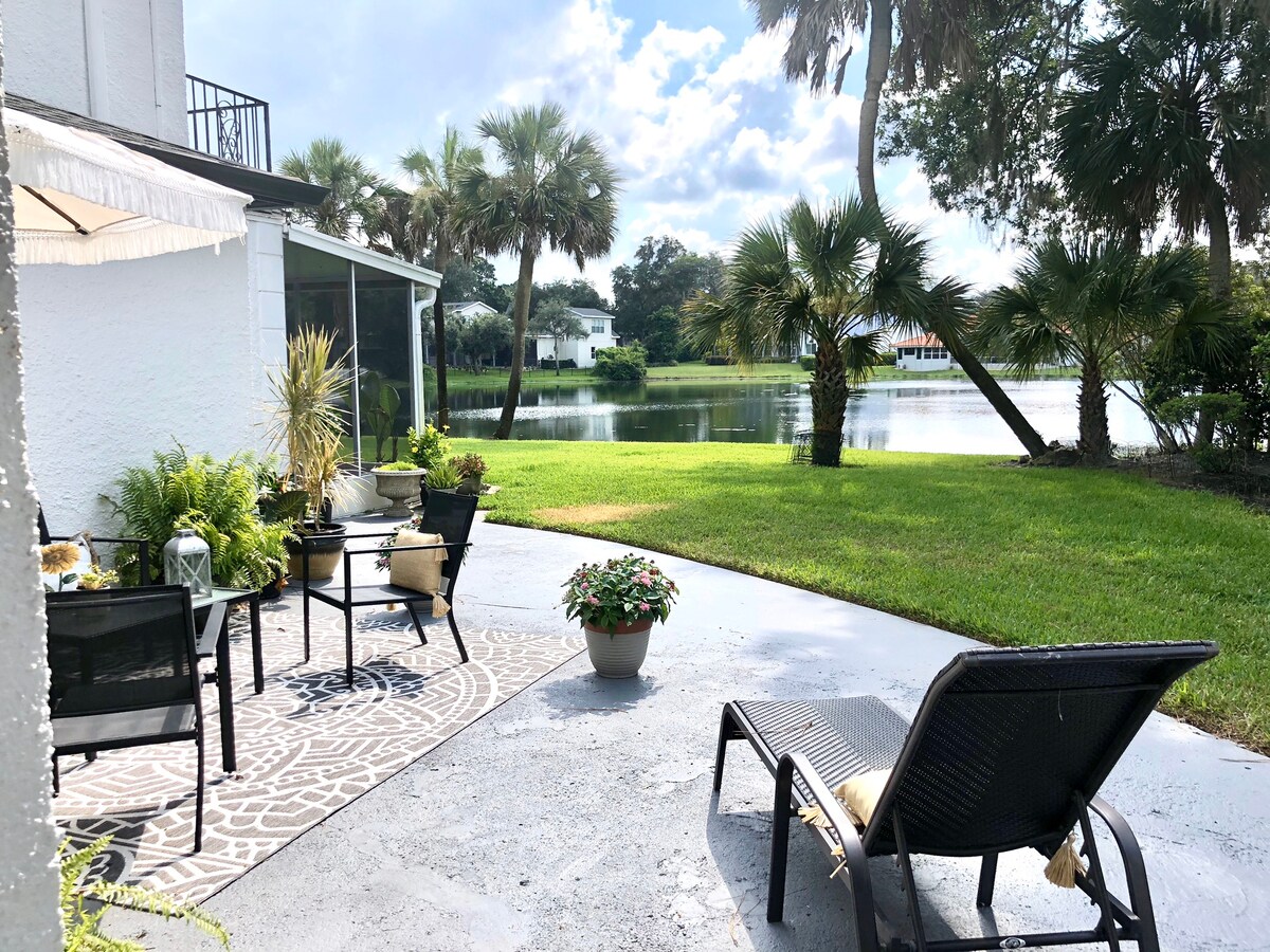 A serene outdoor patio area is visible, featuring black lounge chairs and a patterned rug. Potted plants add greenery, while palm trees frame the scene. A tranquil lake reflects the blue sky and scattered clouds, enhancing the peaceful ambiance of the space.