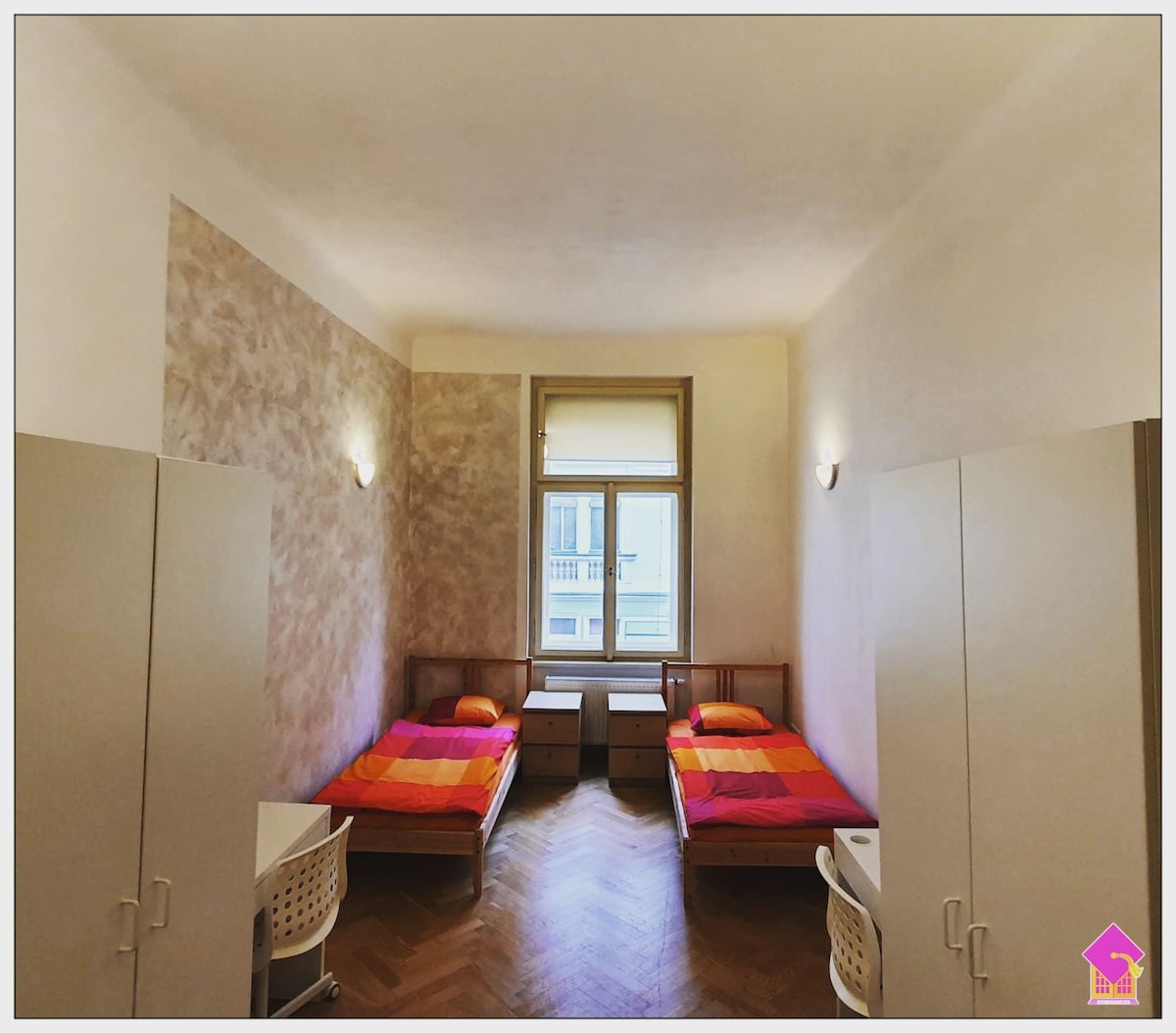 The room features two spacious beds, each with bright orange and pink bedding. Two modern white dressers are positioned beside the beds, complemented by a small desk and chair. Natural light enters through a window, highlighting the warm-toned walls and polished wooden floor.