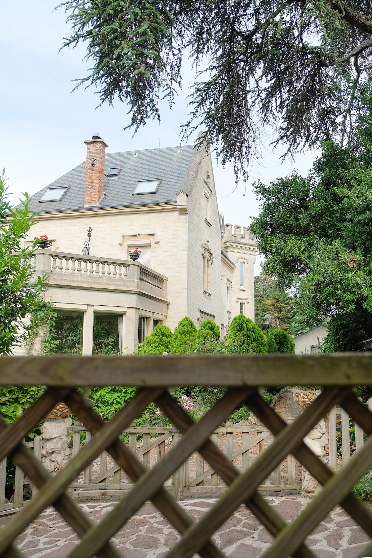 The historic property is framed by lush greenery, showcasing its elegant architectural features. The exterior includes a prominent tower and a stone facade, complemented by a balcony adorned with flowers. Access to the garden is visible, providing a serene outdoor space.