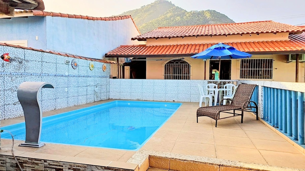 An outdoor pool area features a rectangular swimming pool with a turquoise surface, accompanied by a lounge chair and an umbrella. Light-colored tiled flooring surrounds the pool, while a mountain view and a residential structure are visible in the background.