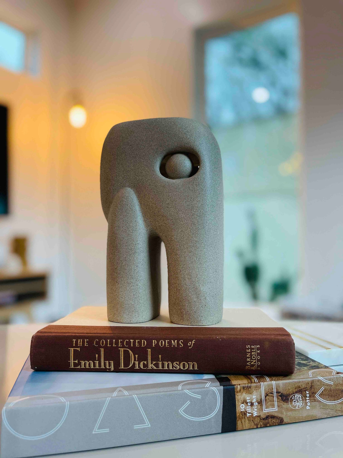 A decorative sculpture stands on top of two stacked books, one featuring the collected poems of Emily Dickinson. Soft light fills the background, subtly highlighting the smooth texture of the sculpture and the titles on the books.