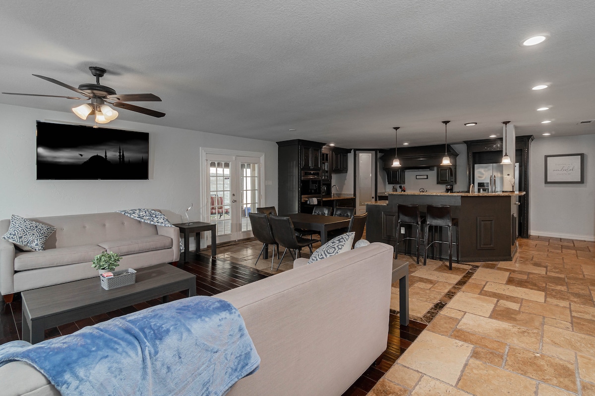 An open-concept living area is displayed, featuring a light-colored sofa set and hardwood flooring. Natural light enters through the glass doors, while a modern kitchen with dark cabinetry and bar seating is visible in the background. A decorative plant adds a touch of greenery.