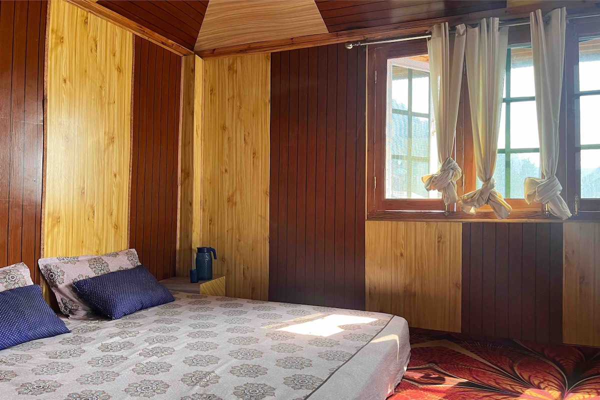 A spacious bedroom is presented with wooden wall paneling and a patterned light-colored bedding. Two large windows allow natural light to fill the room, with soft curtains framing the views outside. A water bottle is placed near the bed, enhancing convenience.
