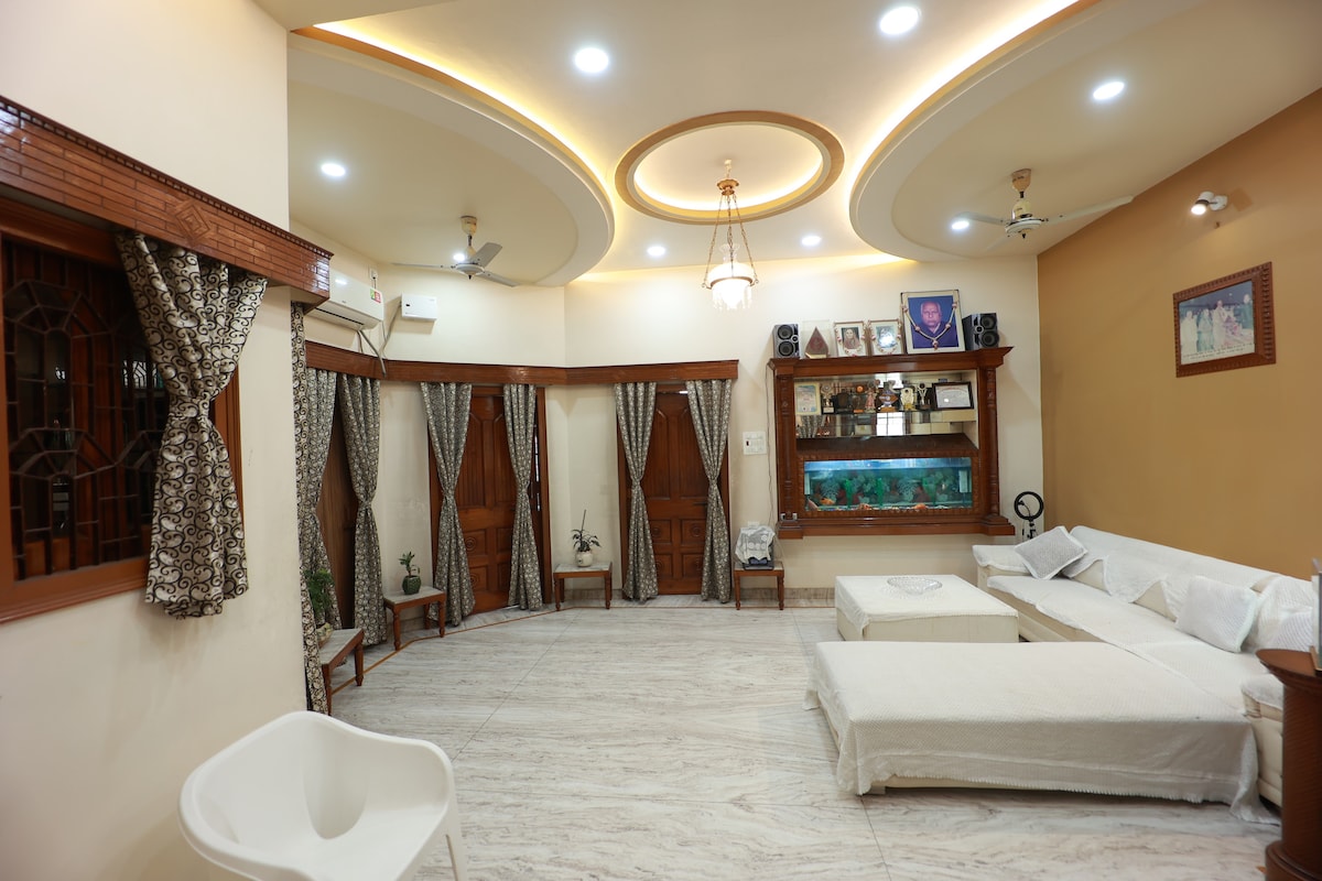 A spacious living area is illuminated by recessed ceiling lights. Large windows with patterned drapes provide natural light, while an aquarium adds a calming touch. Soft seating is arranged around the room, creating an open and inviting atmosphere suitable for relaxation and socializing.