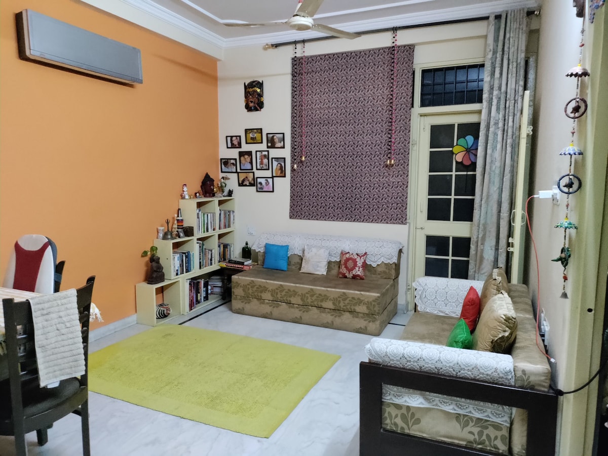 A cozy living area features an orange accent wall, complemented by a plush green rug on the marble floor. A sofa and matching loveseat offer comfortable seating, with decorative cushions adding color. A small bookshelf is filled with books, and framed photos adorn the wall.
