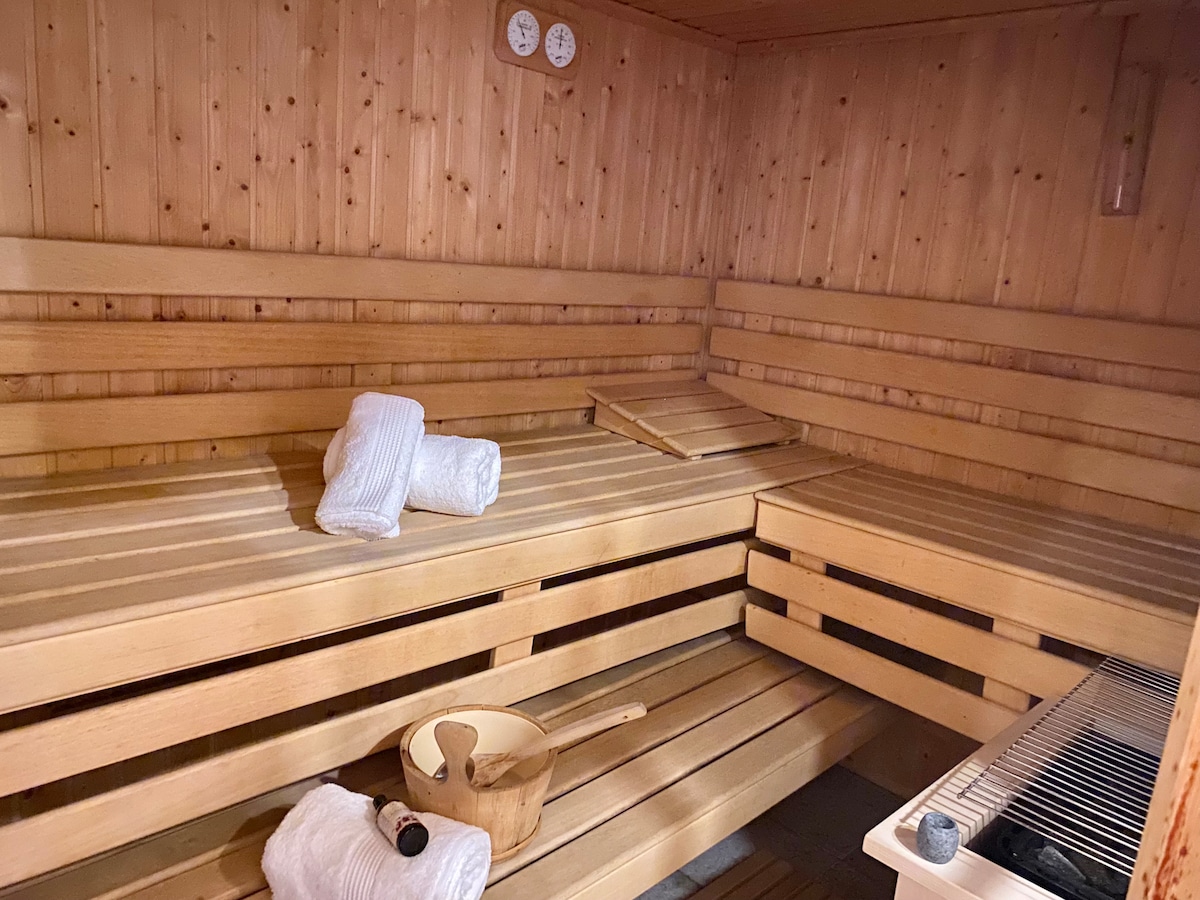 A wooden sauna space is presented, featuring tiered wooden benches arranged along the walls. Soft white towels are neatly placed on the benches. A wooden bucket and ladle are positioned on the floor, while temperature gauges are mounted on the wall.