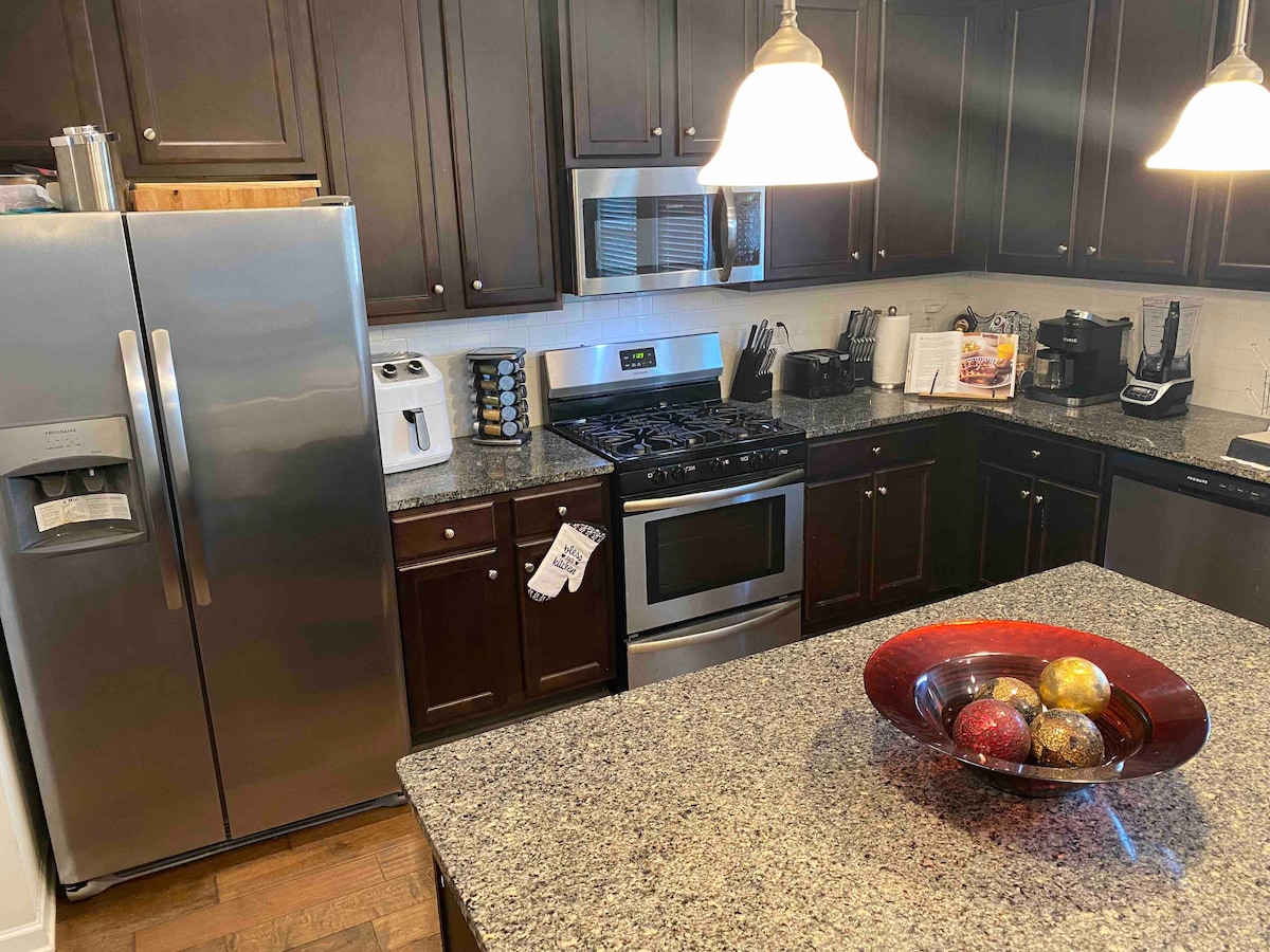 A modern kitchen features dark wood cabinets and stainless steel appliances. A large granite countertop showcases a decorative bowl filled with colorful fruits. The space is well-lit by pendant lights above, and various kitchen tools and appliances are neatly arranged.