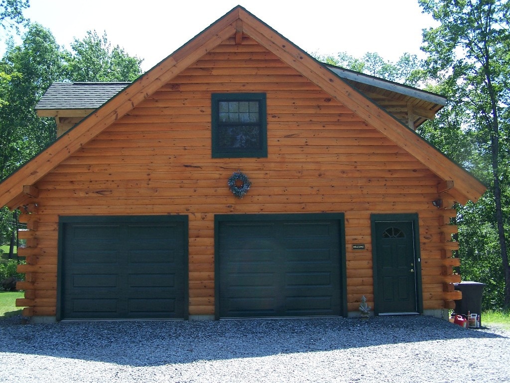 The exterior of a log cabin-style garage is visible, featuring three large doors painted in dark green and a welcoming front door. A gravel driveway leads up to the structure, surrounded by greenery, offering a rustic and inviting appearance.