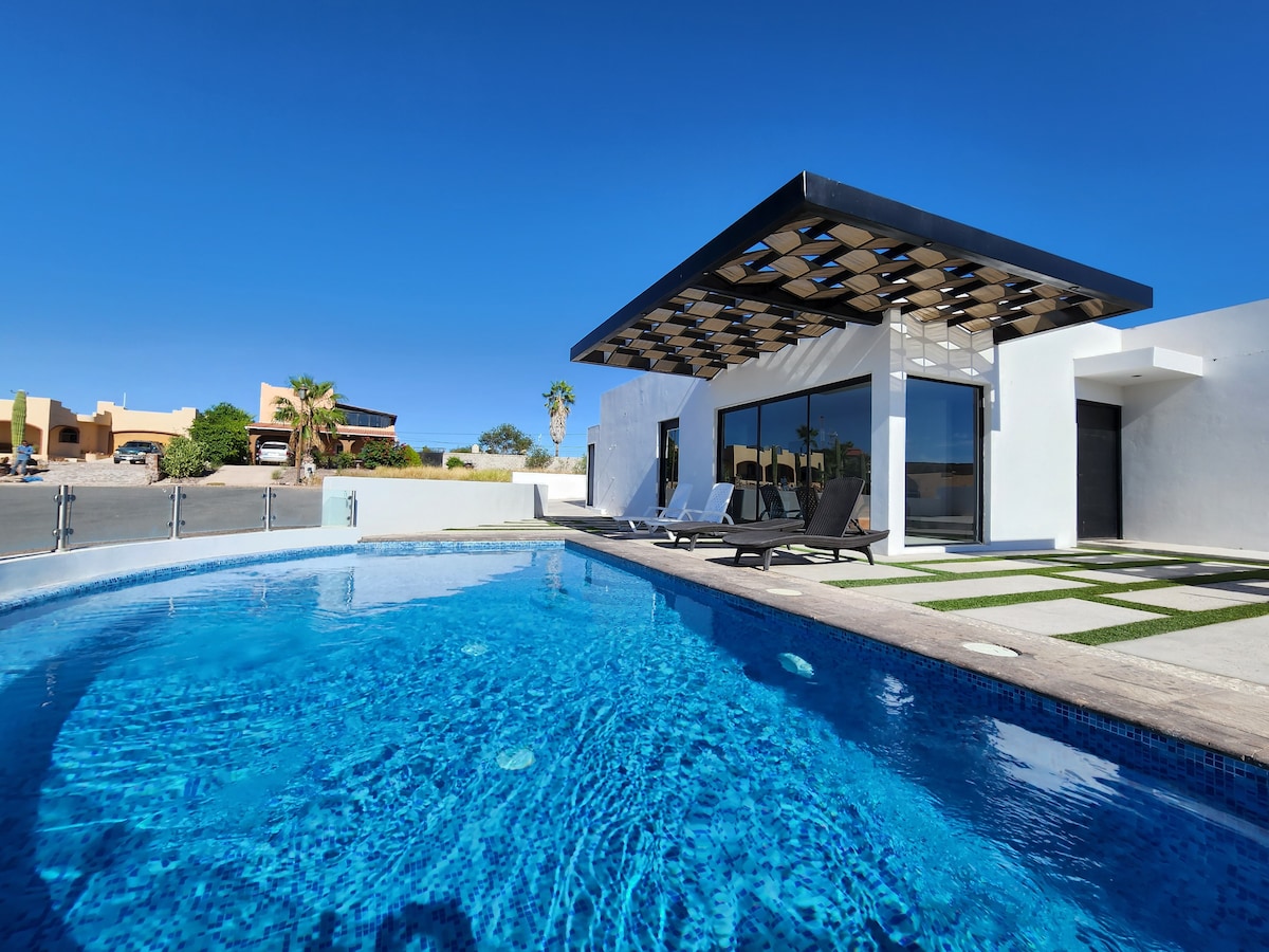A modern house is positioned beside a private pool, featuring a sleek design and a shaded patio. Lounge chairs are placed along the poolside, creating a relaxing space. The landscape includes a clear blue sky, contributing to a serene outdoor setting.