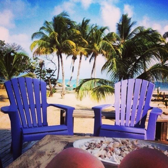 Two purple Adirondack chairs are positioned on a wooden deck overlooking a sandy beach and the ocean. Palm trees sway gently in the background, while a plate of shells rests on a table, capturing the essence of a serene coastal environment.