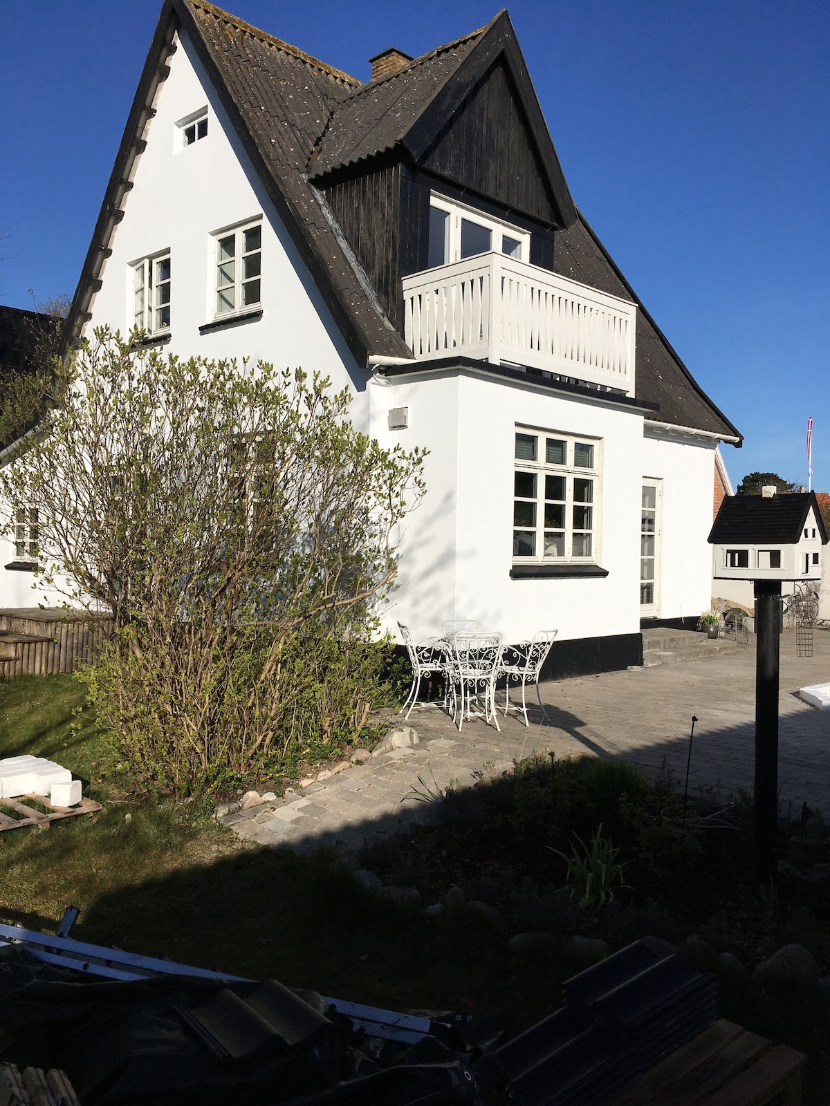 The exterior of the house is depicted, featuring a combination of black and white siding. A small balcony is visible above a set of large windows. A stone pathway leads to a seating area with white metal furniture, surrounded by greenery and a well-maintained landscape.