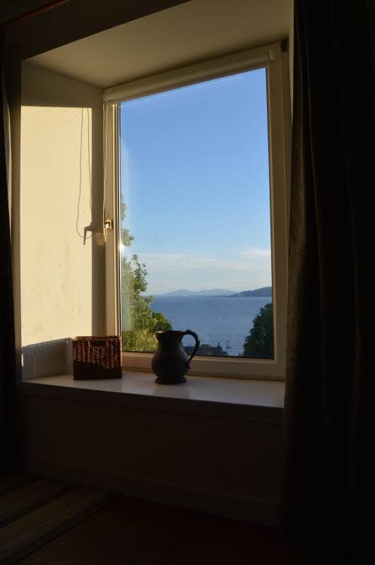 A window offers a scenic view overlooking the water, framed by soft curtains. A decorative jug and a basket rest on the windowsill, complementing the tranquil landscape visible beyond, with distant hills and a calm body of water in the background.