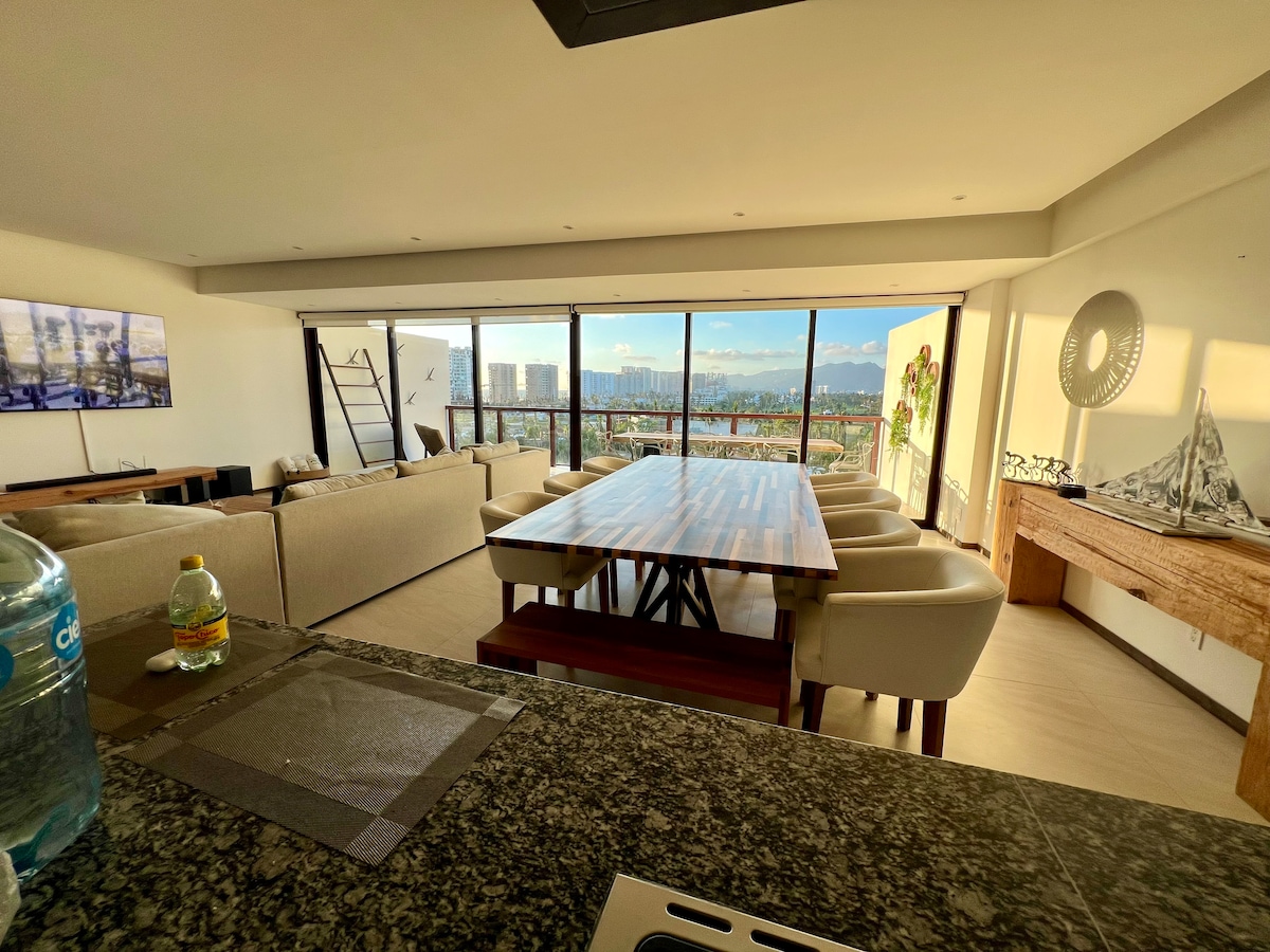 A spacious dining area features a long wooden table surrounded by comfortable chairs. Large windows offer expansive views of the outside landscape, seamlessly connecting the interior with the outdoors. Soft natural light fills the room, emphasizing the modern design and neutral color palette.