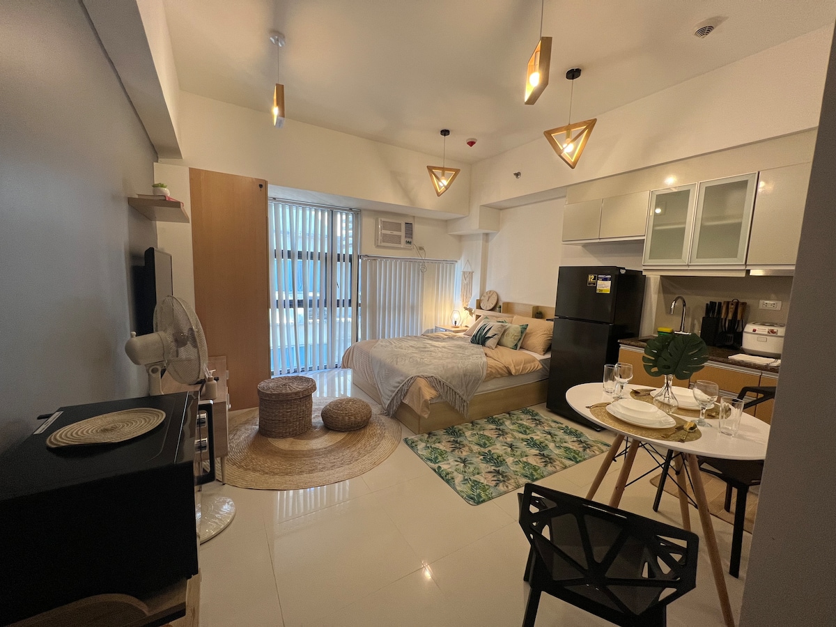 A studio unit features a spacious queen size bed with soft linens, complemented by natural light from the large windows. A dining table, equipped for dining, is set nearby. Gray and neutral tones are used throughout, enhancing the modern aesthetic.
