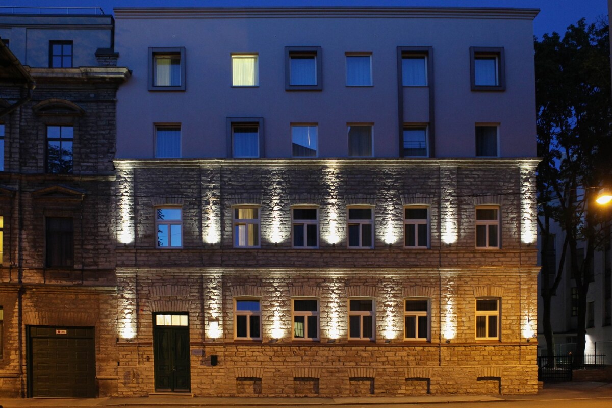 The exterior of the building is highlighted by soft lighting, showcasing a blend of stone and modern architectural features. Windows are evenly spaced along the facade, with a contrasting upper level. The overall design reflects a harmonious mix of historical and contemporary styles.