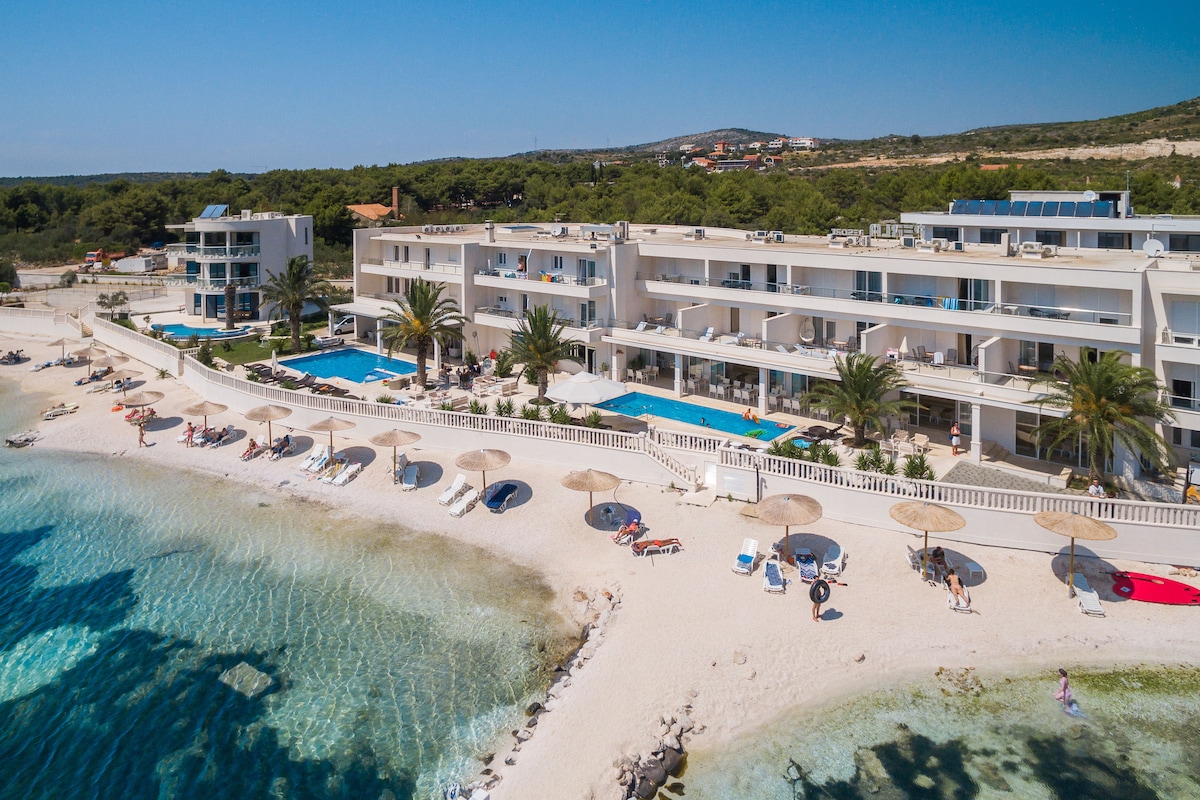 The resort sits by a sandy beach, featuring multiple sun loungers and umbrellas arranged along the shore. A large outdoor pool is offered in front of the building, surrounded by palm trees. The backdrop includes a clear blue sky and coastal greenery.