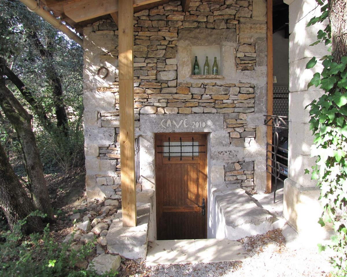 A rustic stone entrance is framed by wooden beams, leading to a cellar. Glass bottles are displayed on a shelf above the door, which features a small, decorative window. Lush greenery surrounds the structure, enhancing its natural setting.