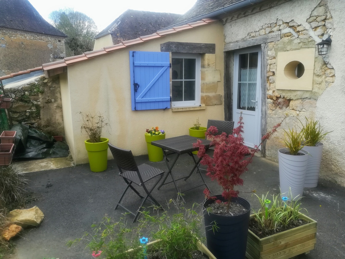 An outdoor seating area features a small, dark table surrounded by four matching chairs. Decorative planters in various colors hold plants, enhancing the space. A blue shuttered window adds a pop of color against the neutral wall of the building.