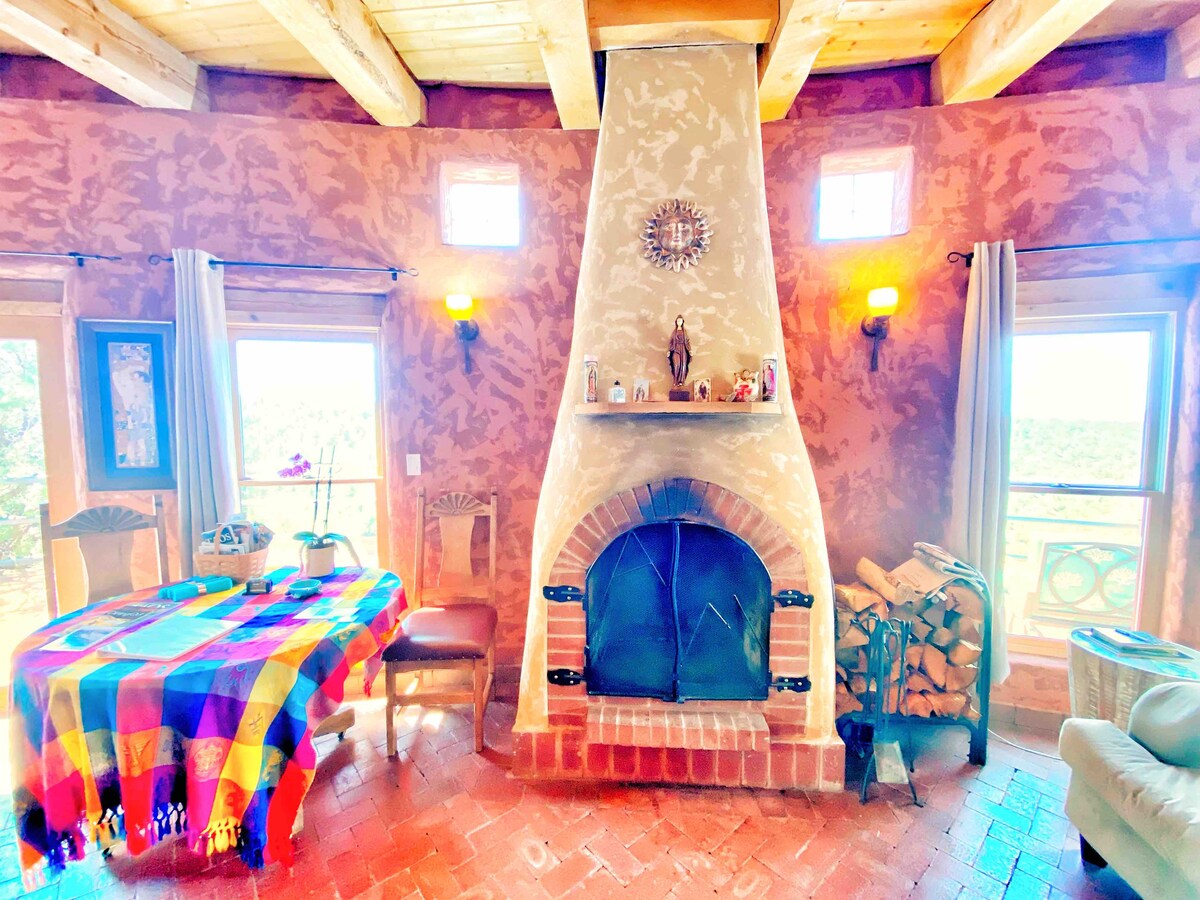 An inviting living area features a large kiva fireplace as the focal point, surrounded by adobe-colored walls. A colorful woven blanket adorns the dining table, while wooden furniture complements the warm atmosphere. Natural light floods in through windows, showcasing scenic views outside.