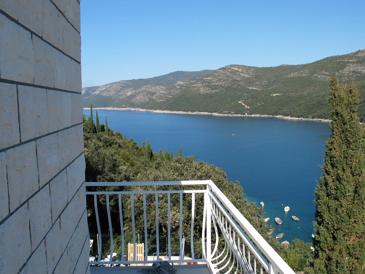 A scenic view showcases calm waters reflecting the clear blue sky, framed by lush greenery and distant hills. The image captures a tranquil coastal landscape with boats nestled in the bay below, enhancing the serenity of the environment.