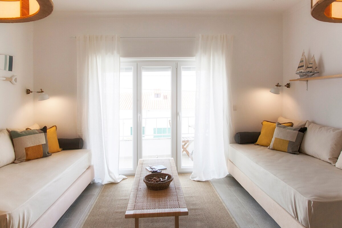 A light-filled living area features two spacious daybeds adorned with neutral and warm-toned pillows. Soft curtains frame the large window, leading to a small balcony. The natural fiber rug adds texture to the space, complemented by two ceiling fixtures that enhance the inviting feel.