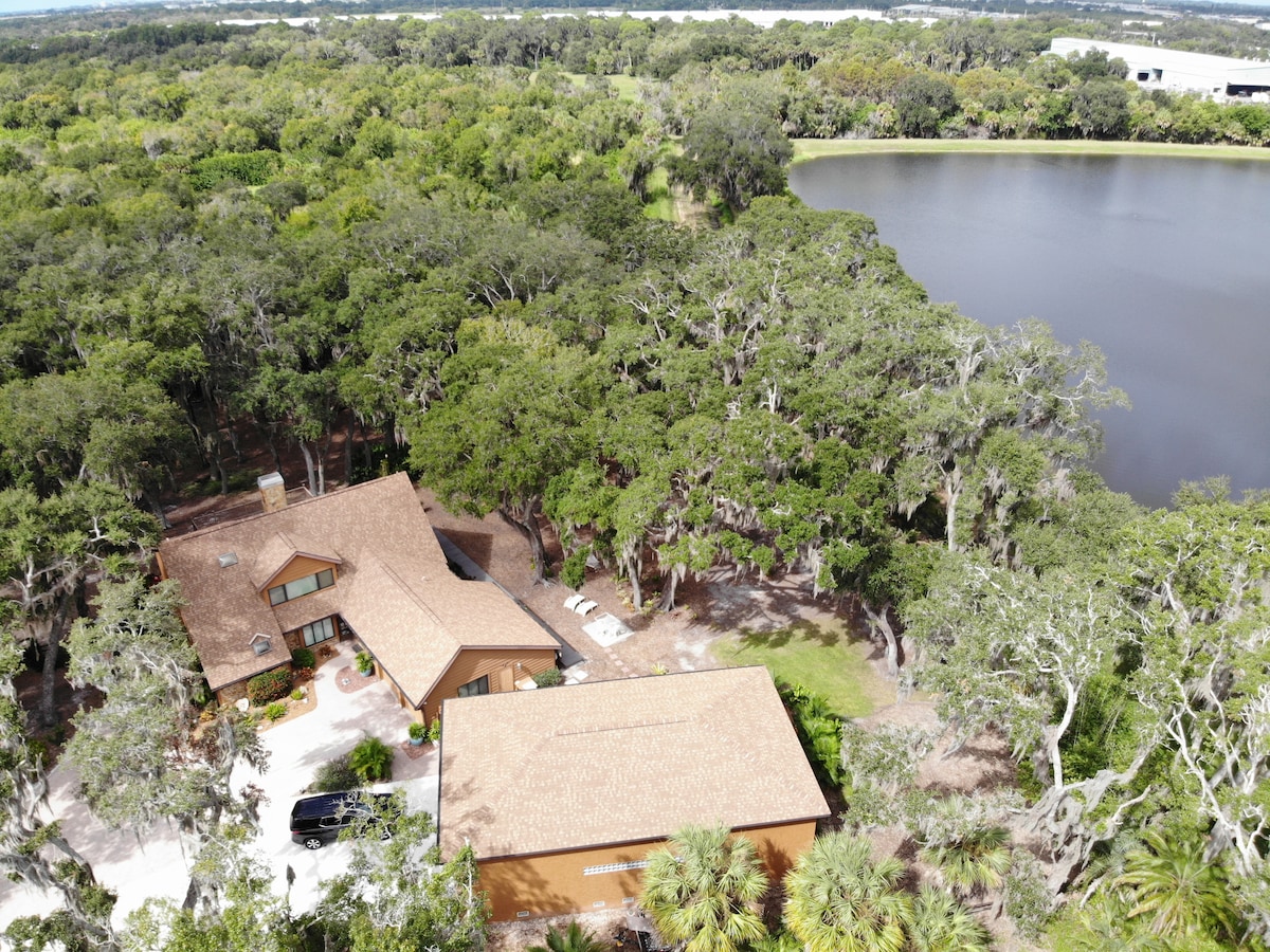 An aerial view showcases a large detached building surrounded by mature oak trees and lush greenery, with a serene lake visible in the background. The expansive yard complements the peaceful setting, offering ample space for outdoor activities.