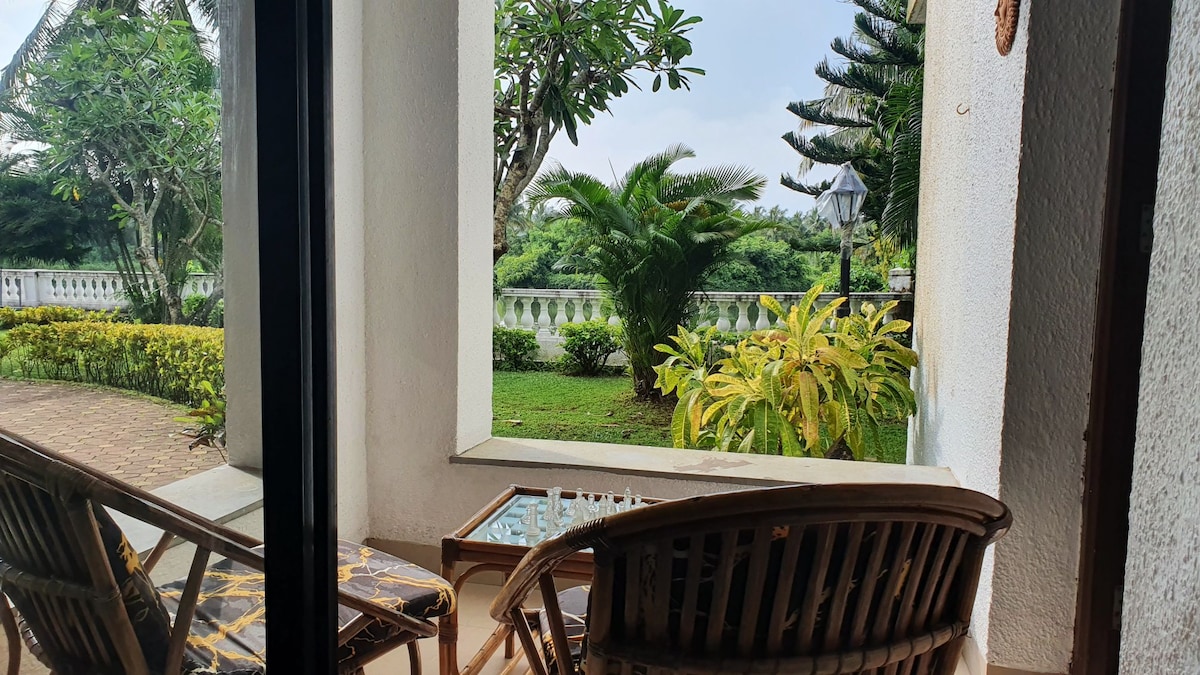 A serene outdoor seating area is visible, featuring two wooden chairs positioned on a patio. Lush greenery and tropical plants frame the view, while a chessboard sits atop a small table. The ambiance suggests a relaxing space for enjoying the natural surroundings.