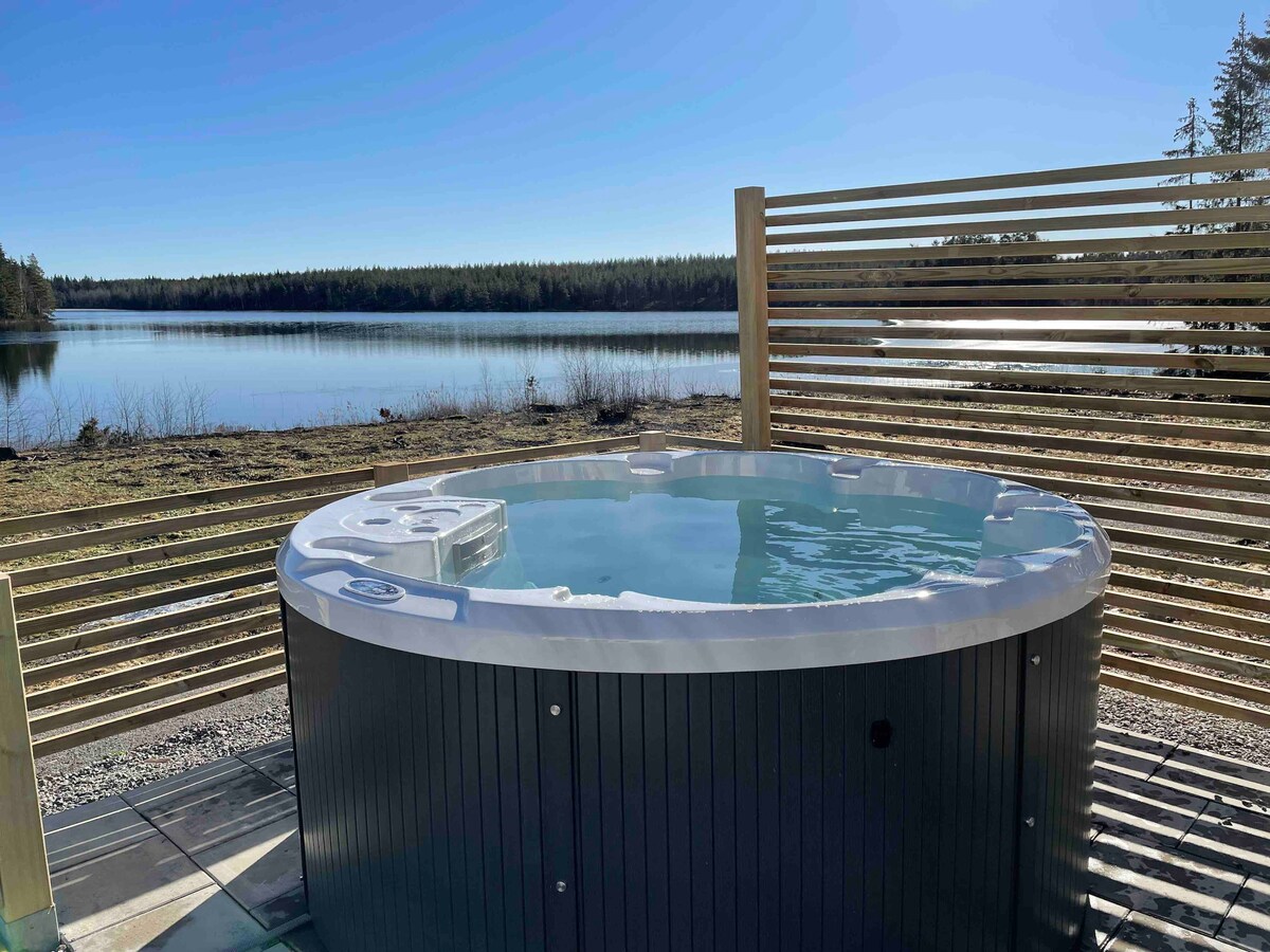 A warm, electric heated hot tub is positioned outdoors, surrounded by a wooden slatted screen for privacy. The serene water reflects the clear blue sky and offers views of the lake and trees in the background.