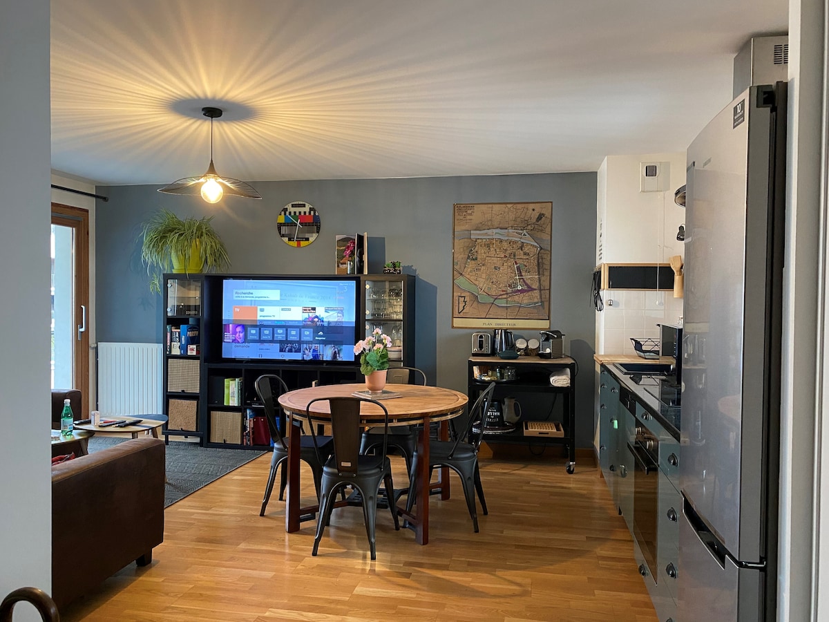A modern kitchen and dining area is visible, featuring a round wooden table surrounded by black chairs. A large screen provides entertainment options. Shelving displays various decorative items, and kitchen appliances are neatly arranged along the counters.