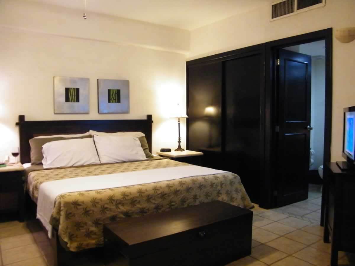 A spacious bedroom features a large bed with light bedding and decorative pillows. Family photos are placed on a small bedside table beside a lamp. A dark wooden dresser and a television are present, while framed art adorns the walls. A door leads to an adjoining area.