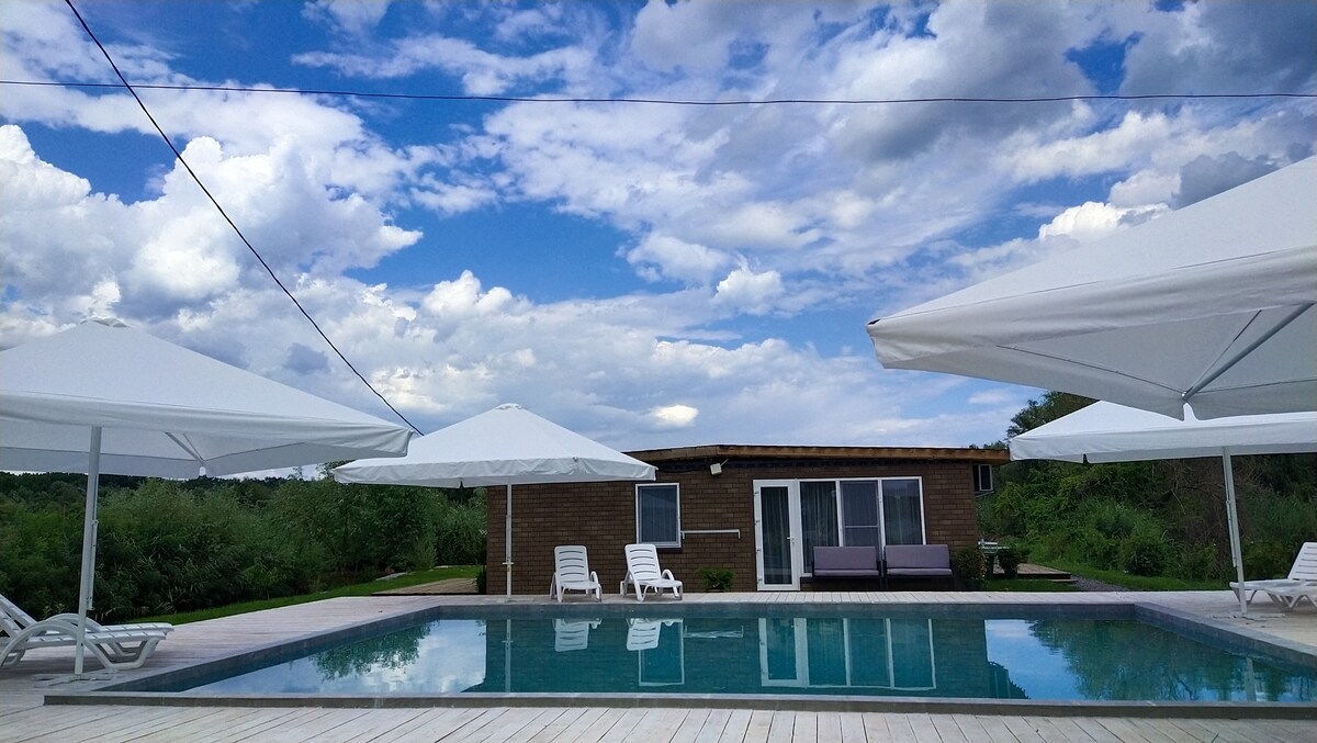 A spacious outdoor area features a tranquil swimming pool surrounded by shaded lounges and umbrellas. The pool reflects the sky, creating a serene atmosphere. In the background, a cottage with large windows complements the natural setting.