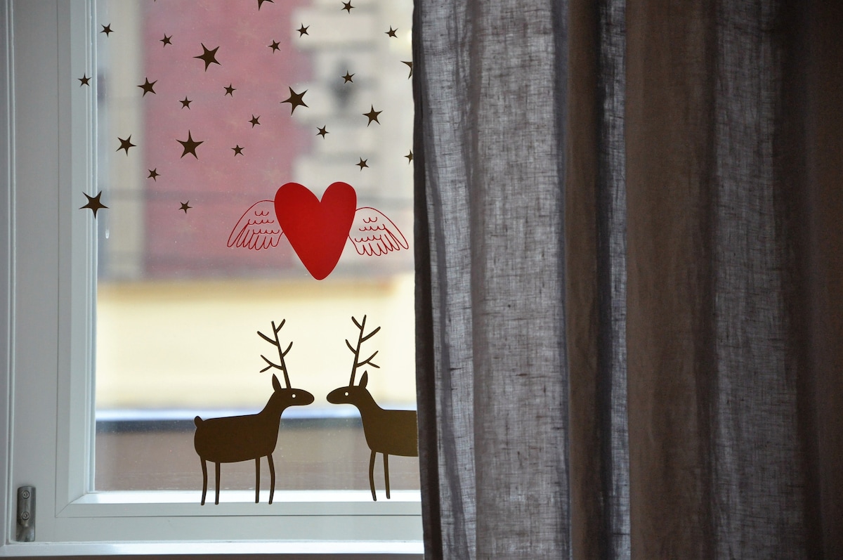 A window adorned with festive decorations features two reindeer with antlers and a heart with wings. Light gray curtains partially block the view, creating a cozy ambiance. The background hints at the historical architecture of the surrounding area.