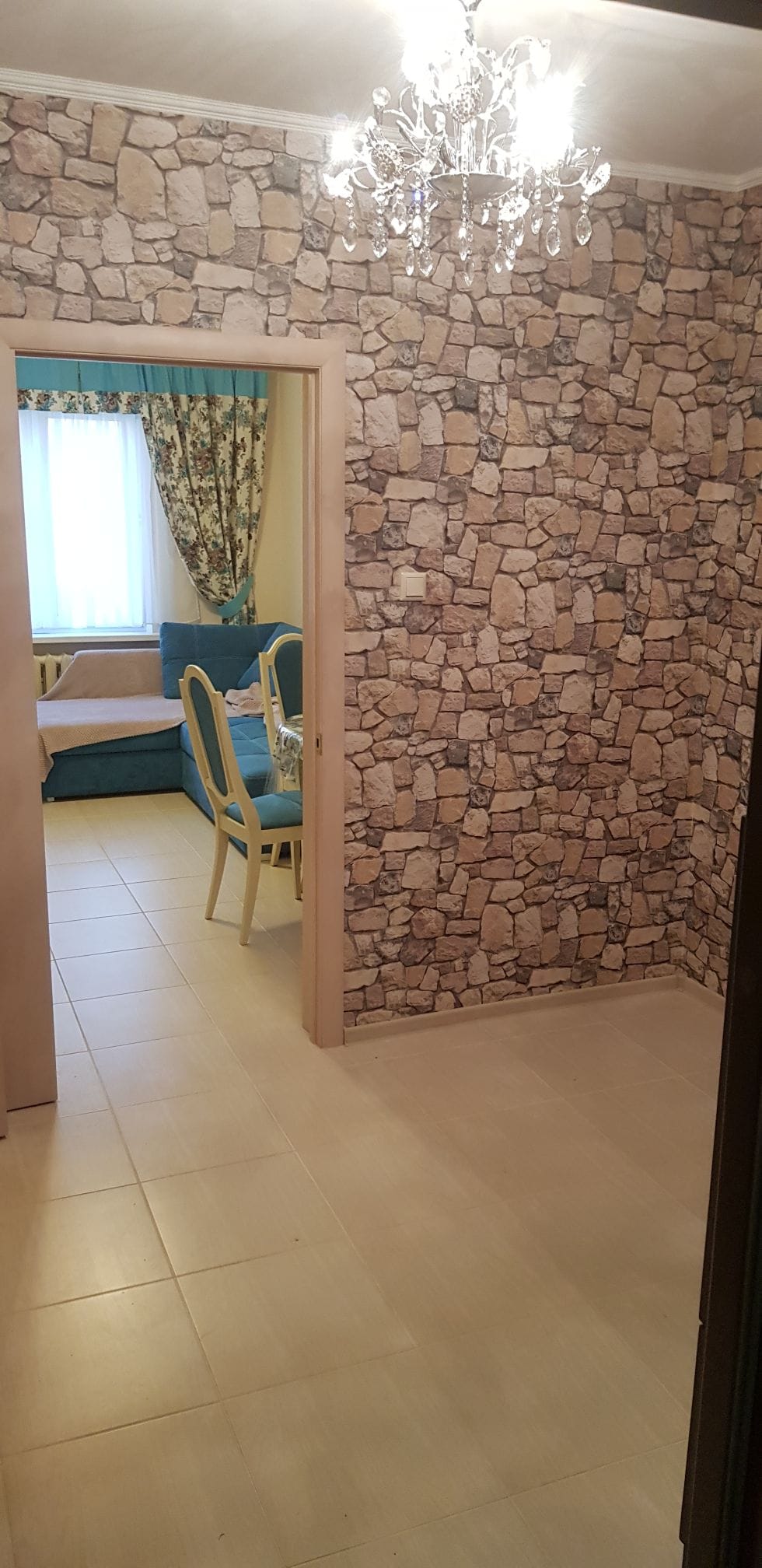 A welcoming entryway is adorned with a stone-patterned wall. A partially visible living area features a sofa in soft hues, accompanied by a dining table and chairs. Natural light filters through a window, enriched by patterned curtains, enhancing the inviting atmosphere.