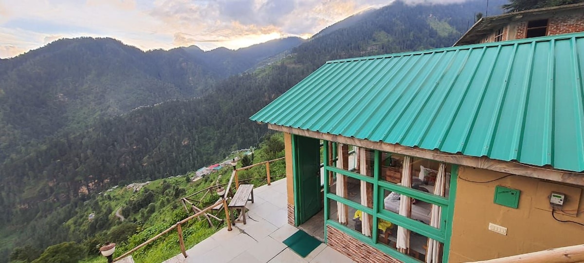 A mountain retreat features a vibrant turquoise roof, surrounded by lush hills. Large windows provide expansive views of the valley below, while an outdoor seating area offers a place to enjoy the fresh air. Stone pathways lead through the greenery.