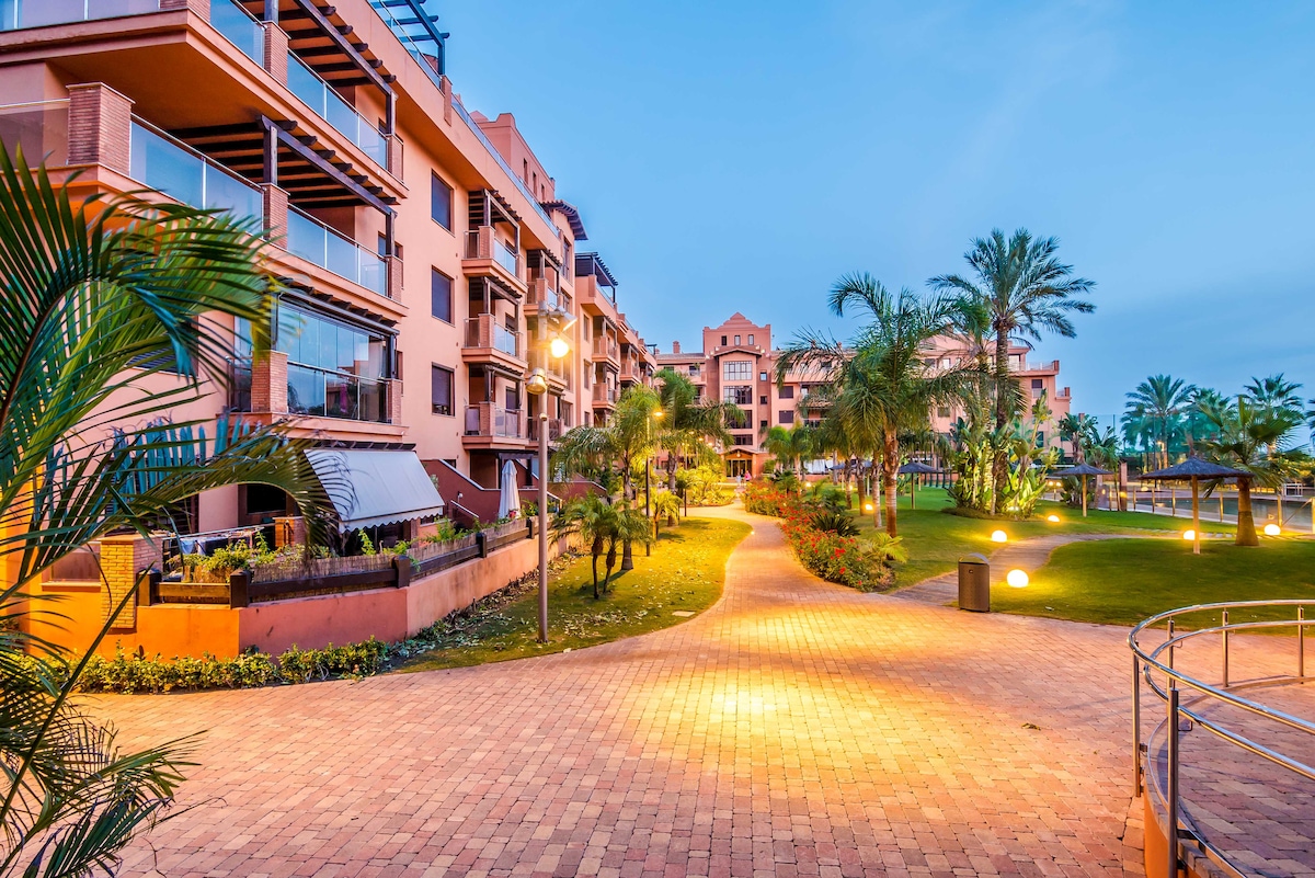 A well-maintained pathway is lined with palm trees and landscaped greenery, leading through a tranquil residential area. Soft lighting enhances the evening ambiance, illuminating the nearby buildings and creating a serene atmosphere for evening strolls.