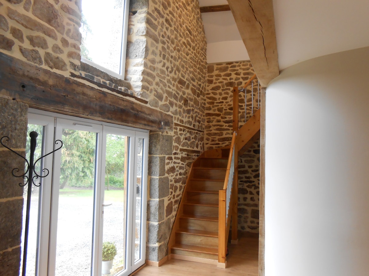 A sunlit entrance hall features a mix of stone and wood elements, showcasing exposed beams. A wooden staircase leads to a spacious living area above. Large glass doors provide views of the outside, inviting natural light into the space.