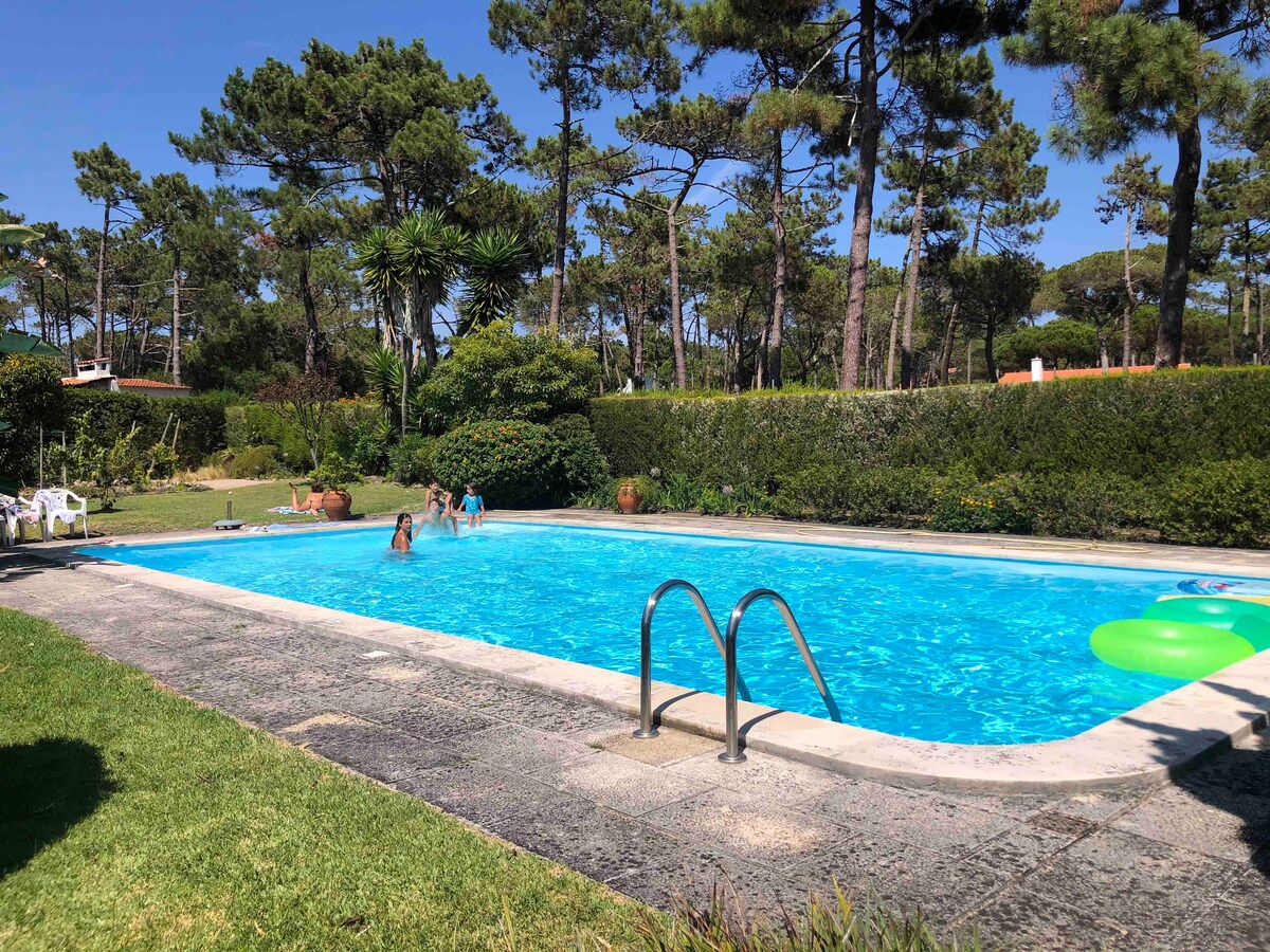 A large outdoor pool is nestled within a well-maintained garden, surrounded by lush greenery and tall trees. Sunbathers can be seen enjoying the water, while colorful floatation devices add a playful touch to the inviting space.