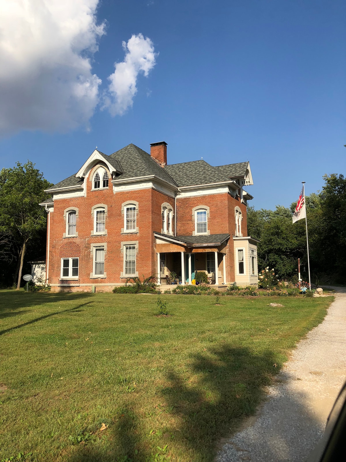 The historic house, built circa 1890, features a striking red brick façade and multiple gabled roofs. Surrounding a manicured lawn, landscaped gardens include various plants. An American flag stands near the entrance, and the pathway leads toward the welcoming front porch.