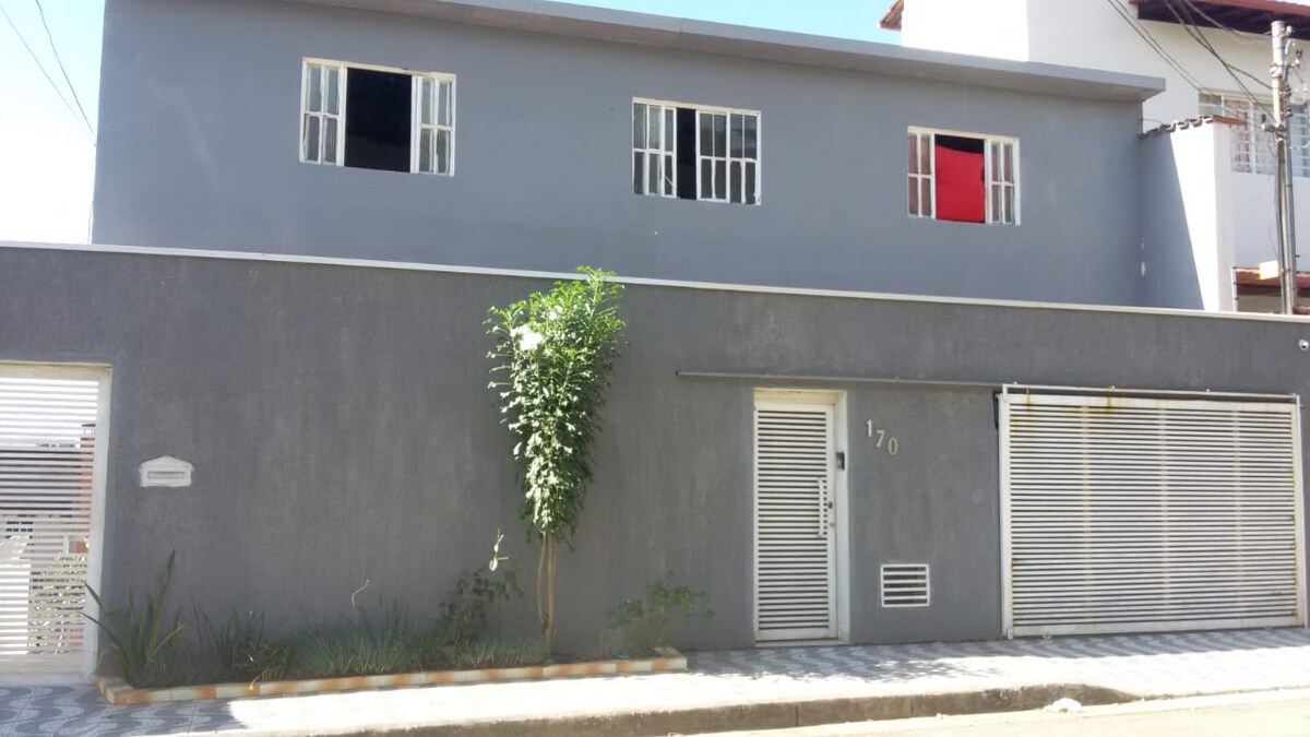 A modern, two-story house features a grey exterior with multiple windows, two of which are open. A small green plant is positioned near the entrance, and a secure gate is visible leading to the front door. The street in front is paved with patterned tiles.