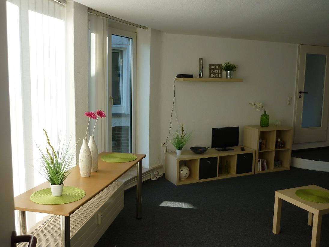A cozy living space is presented with natural light streaming through large windows. A small dining table is set with green placemats, while a shelving unit holds decorative items. A television and additional furnishings contribute to the welcoming atmosphere.