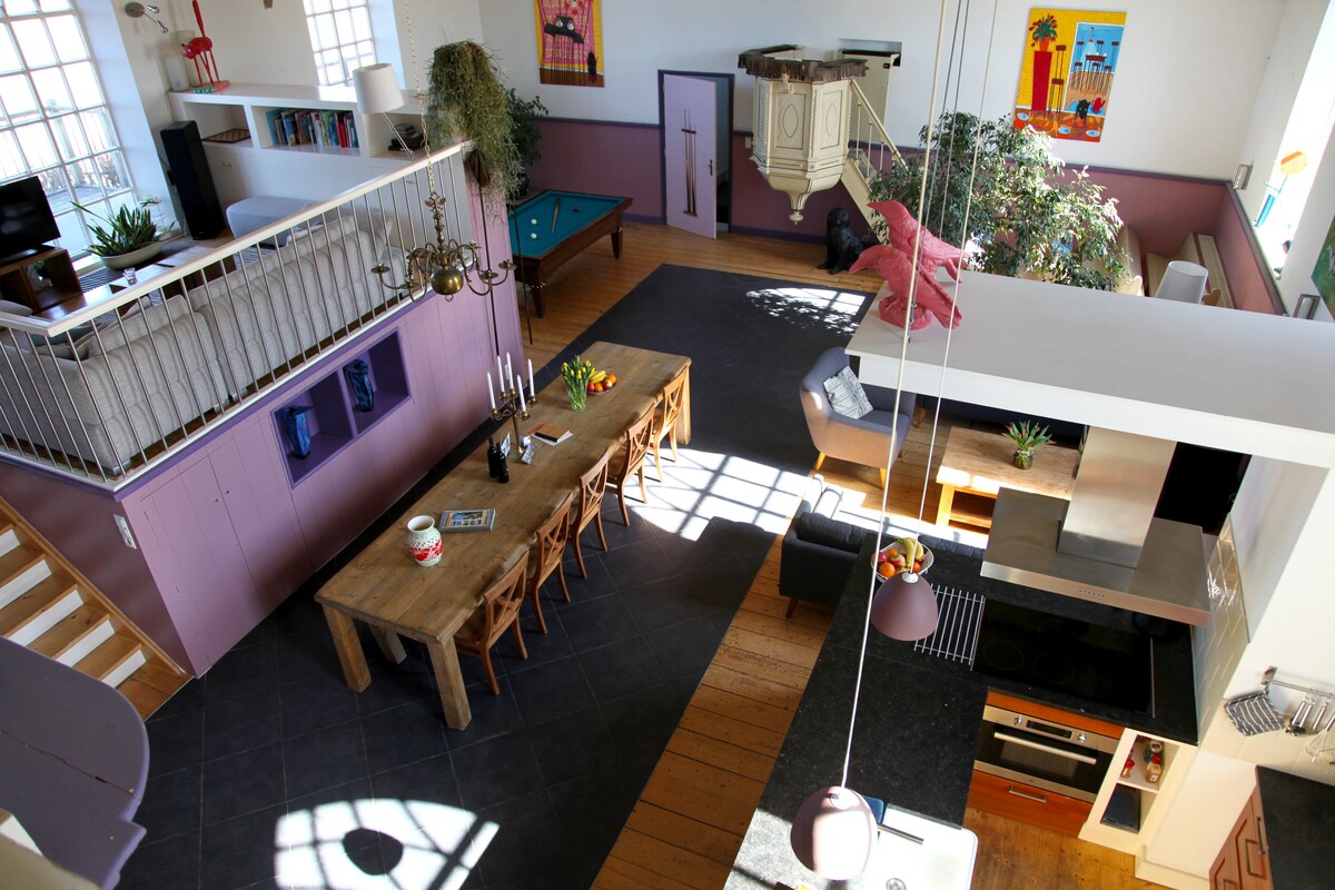 The spacious interior features high ceilings and large windows, allowing natural light to fill the area. A long dining table with chairs is positioned near a well-equipped kitchen. A pool table adds to the recreational options, while decorative plants enhance the open layout.