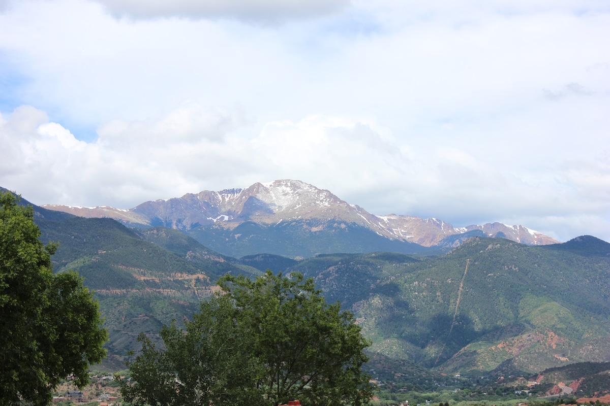 A panoramic view of snow-capped mountains stretches across the horizon, surrounded by verdant hills and trees. Soft, gray clouds drift above the peaks, suggesting a serene and inviting atmosphere in the backdrop of a scenic landscape.