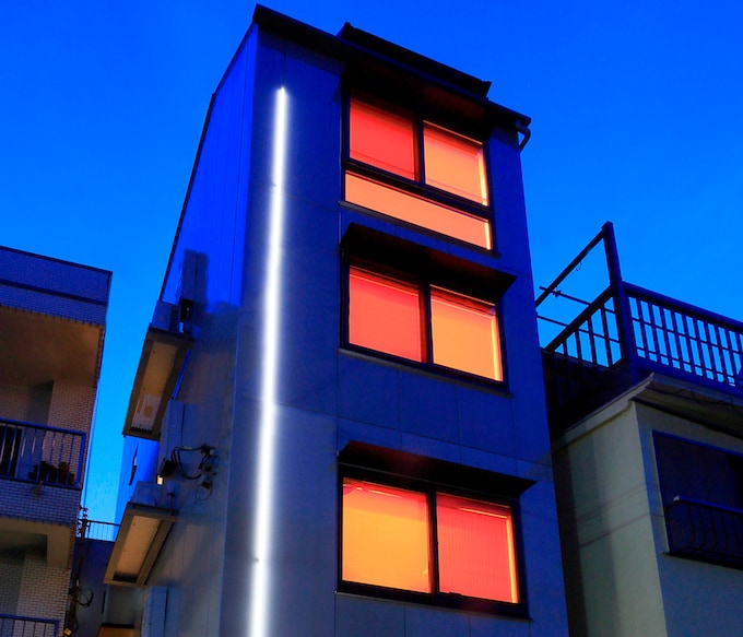 The modern building features multiple floors, characterized by large windows with vibrant orange shades. A sleek, illuminated strip runs along the side, creating a striking visual contrast against the evening sky.