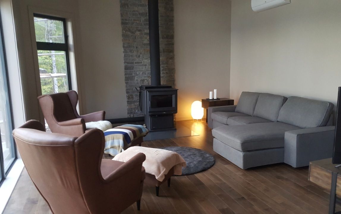 A modern living area features a gray sectional sofa and two brown armchairs, arranged on warm wood flooring. A stone wood stove stands against the wall, accompanied by a circular area rug. Natural light enters through large glass windows, enhancing the open space.